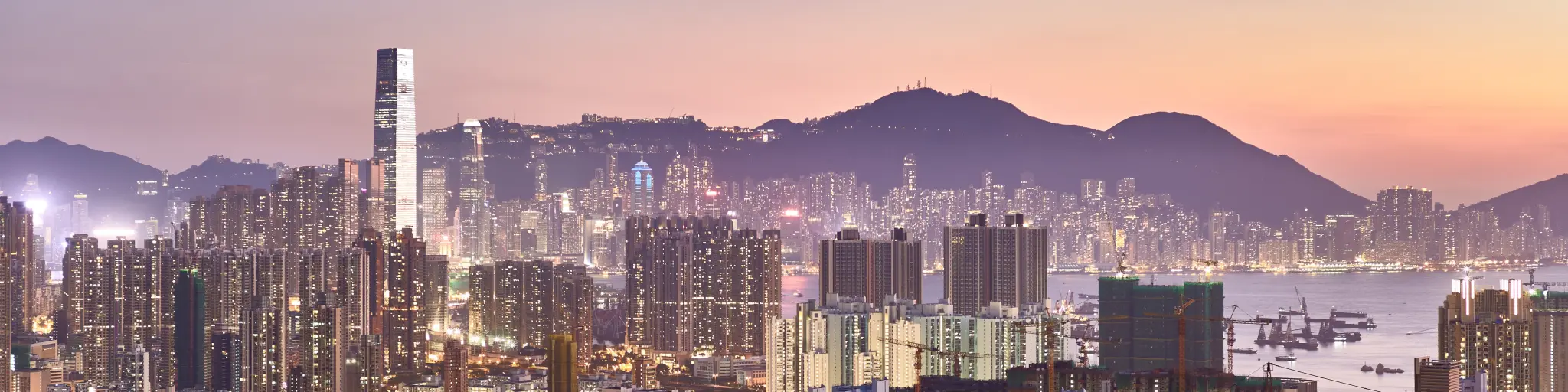 The Hong Kong skyline lit up amid a pink sunset with the harbour in the middle and mountains in the background