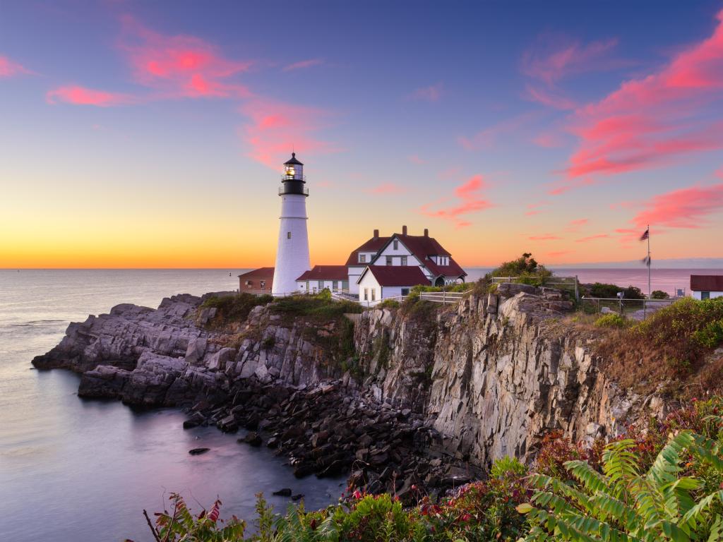 Portland Head Light in Cape Elizabeth, Maine, USA taken at sunset with the lighthouse overlooking the calm sea and rocky cliffs in the foreground. 
