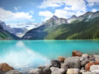 Banff National Park, Alberta, Canada with the stunning turquoise water of Lake Louise in the foreground, with forest covered mountains and snow-capped mountains in the distance on a cloudy but sunny day.