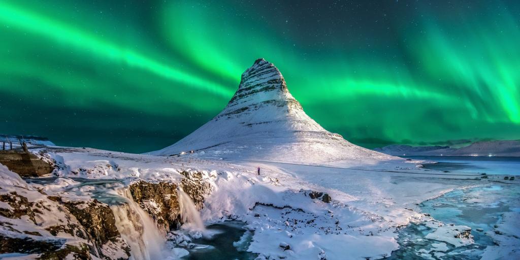 Green Northern lights in the sky over Iceland, with a snow capped mountain and waterfalls below it