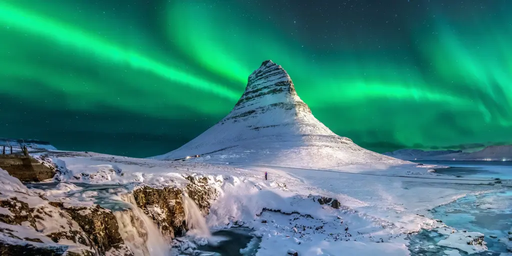 Green Northern lights in the sky over Iceland, with a snow capped mountain and waterfalls below it