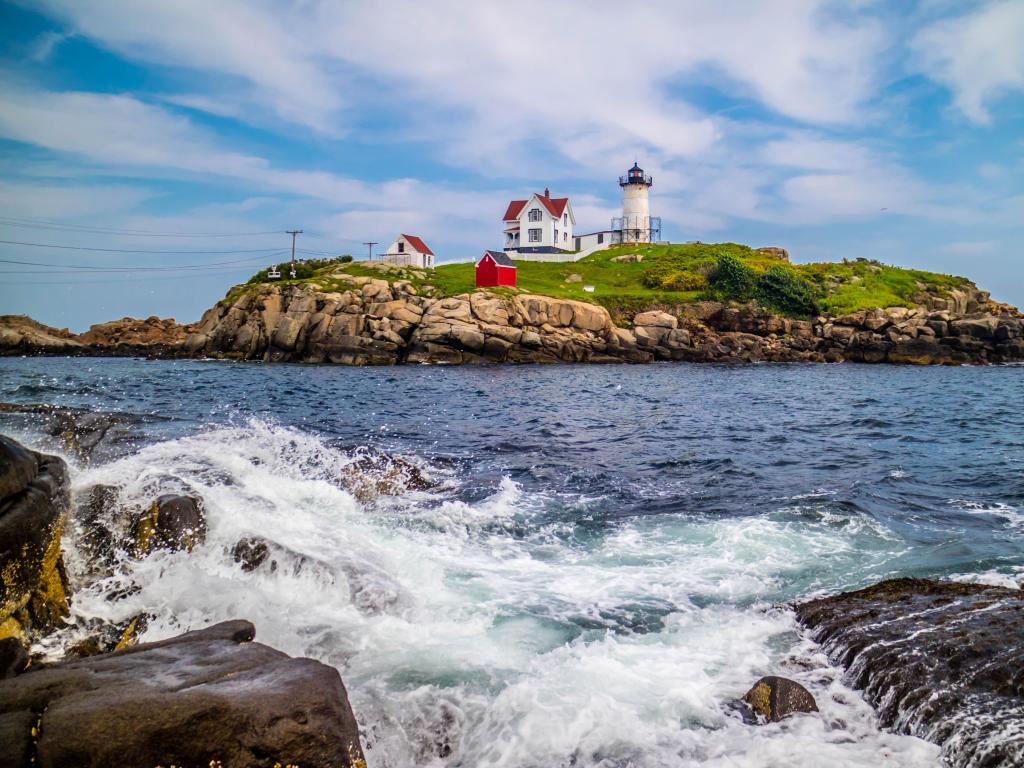Nubble Lighthouse, Maine USA with a sea hitting rocks in the foreground and an island on the horizon with a lighthouse and a few buildings dotted beside it on a cloudy day.