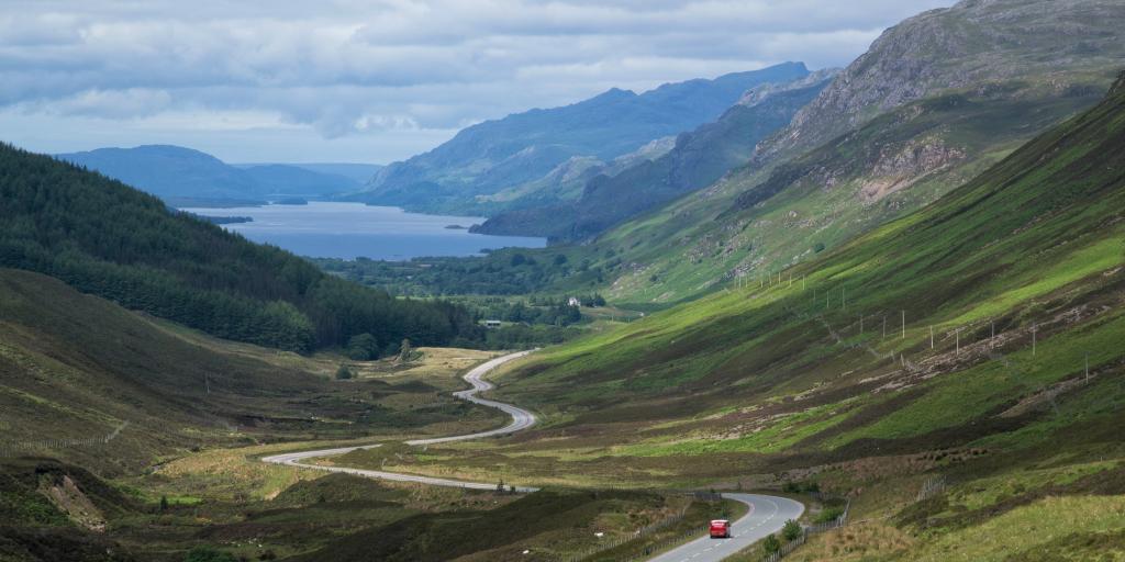 A red car driving down a winding road between two mountains, with a loch in the background