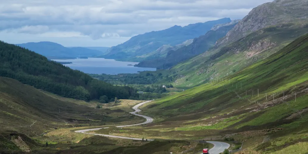 A red car driving down a winding road between two mountains, with a loch in the background