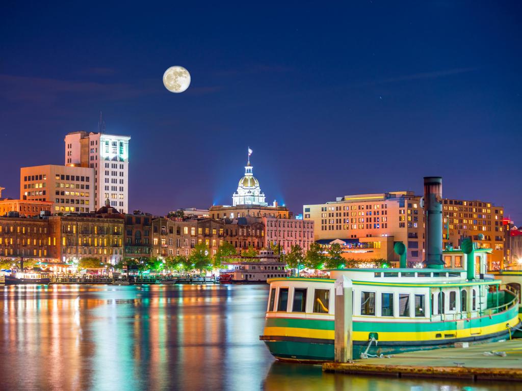 Savannah, Georgia, USA taken at the historic district waterfront of at twilight with a boat in the foreground and the buildings in the distance and the moon above.