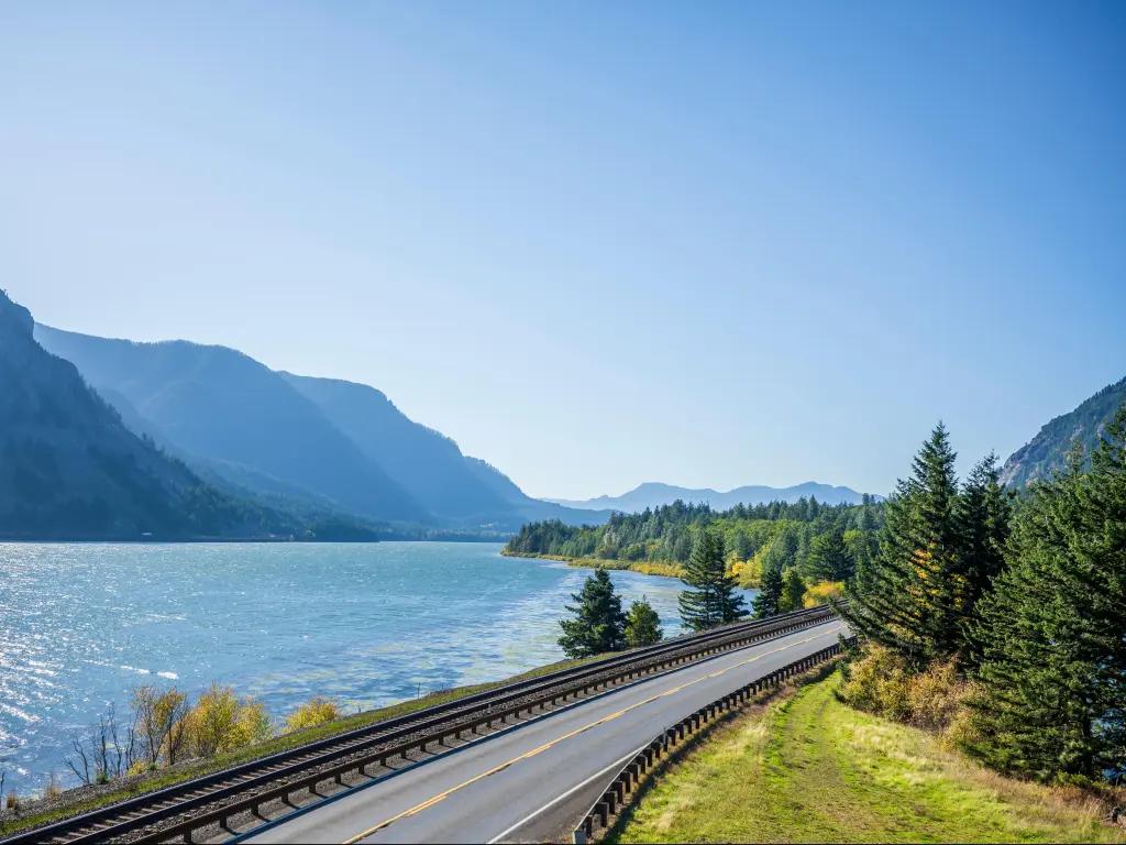 A railroad track next to a highway road with lane dividing opposite traffic runs along the scenic Columbia River bank with green trees and rocky mountains in Columbia Gorge area
