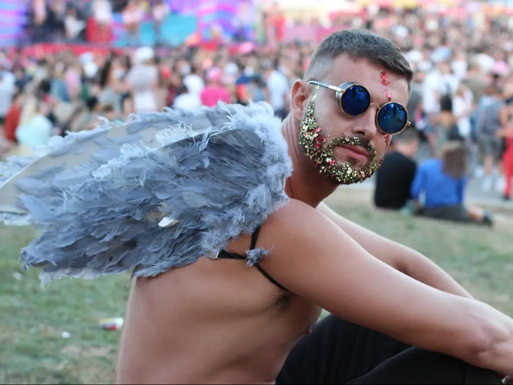 A festival-goer with glitter and angel wings sits and enjoys a music festival