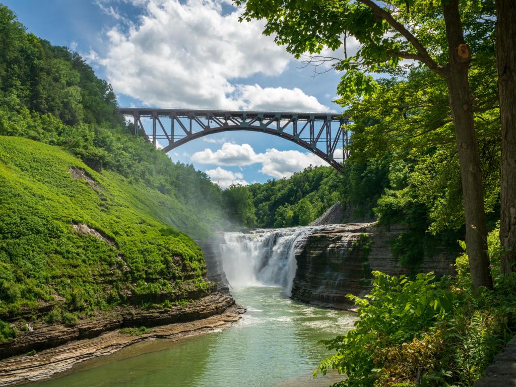 Letchworth State Park, New York, USA with Upper Falls And Genesee Arch Bridge in the background, a waterfall and river in the foreground and surrounded by trees on a sunny day.