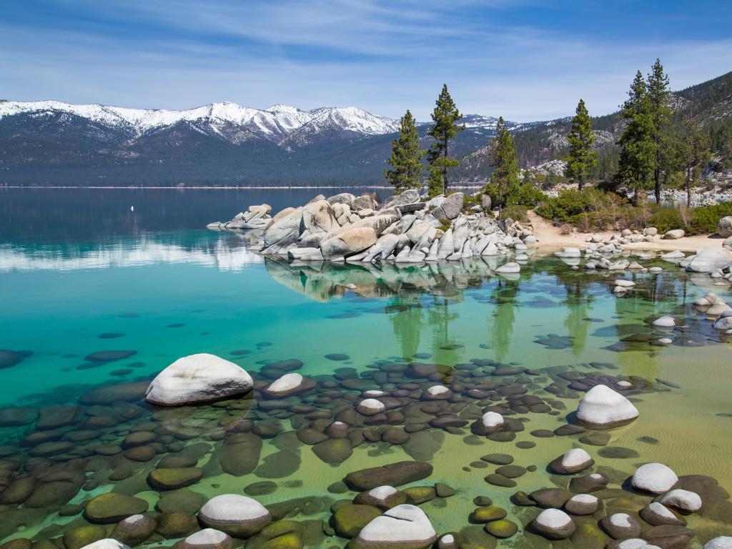Sand Harbor, Lake Tahoe with turquoise clear water and large rocks in the lake, trees and then the snow-capped Sierra Nevada Mountains in the background on a sunny day.