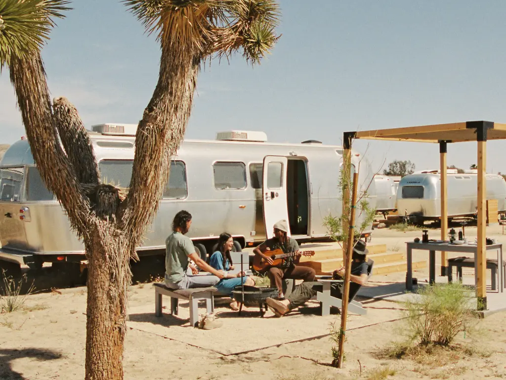 A group of people sitting in front of an Airstream van, one of them is holding a guitar, Joshua Tree in the foreground