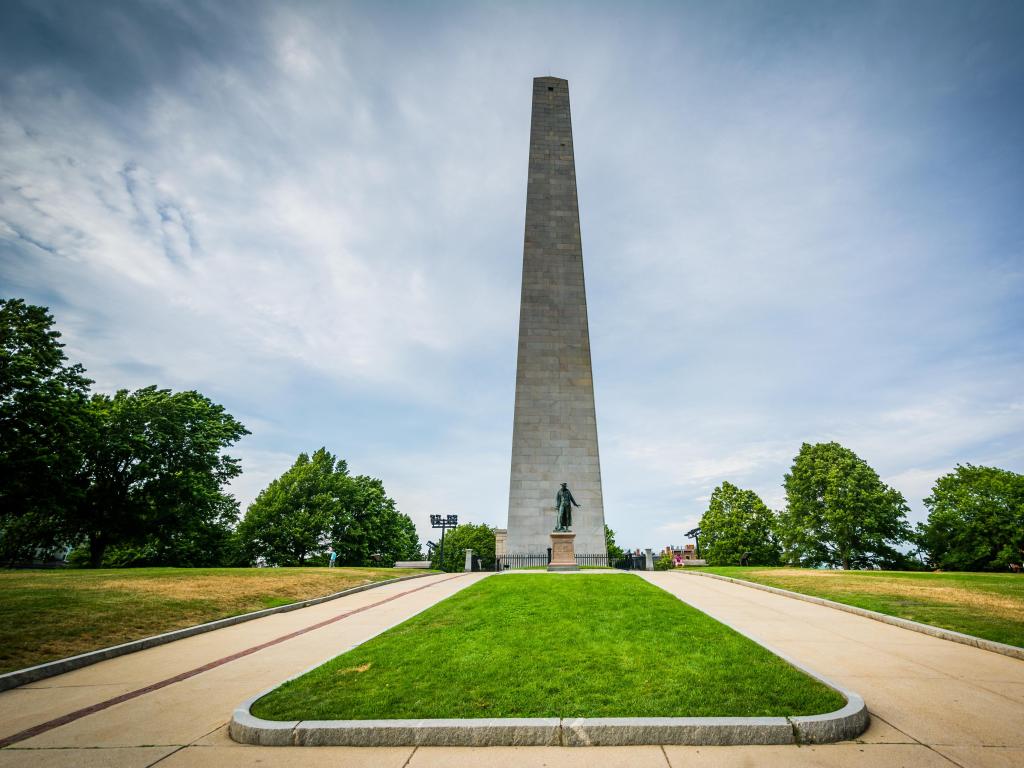 The Bunker Hill Monument, Charlestown, Boston, Massachusetts, USA taken on a sunny day with grass in the foreground and trees in the distance.