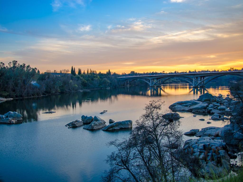 Bridge over wide river at sunset with trees reflected on still water and rocks in the foreground