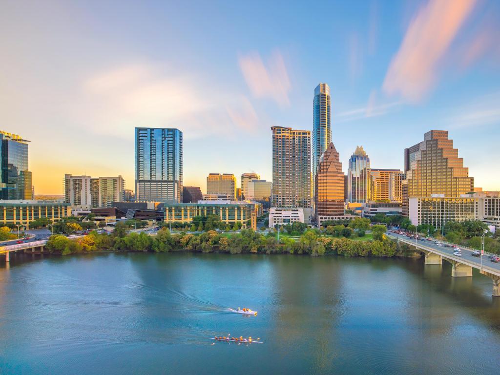 Wide blue river with rowing boat, high rise buildings along river bank with pink and blue sunset sky