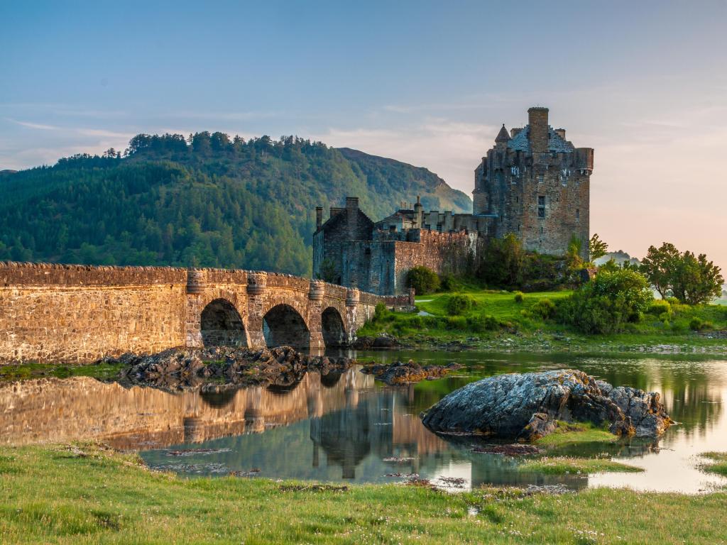 Eilean Donan Castle, Scotland, UK with a river and bridge in the foreground and the castle against a hilly mountain at sunset.