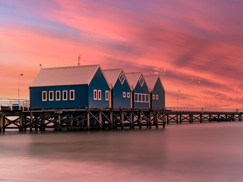 Wooden jetty stretches into the distance over the ocean with three wooden buildings in the foreground, under a vivid pink sunset sky.