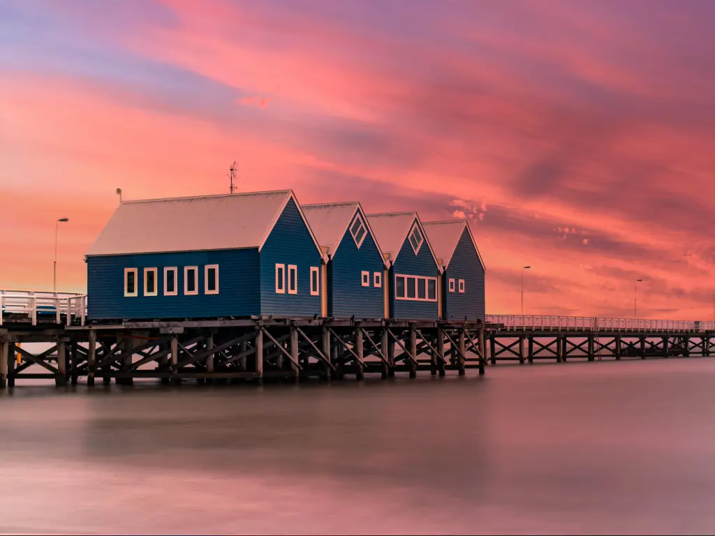 Wooden jetty stretches into the distance over the ocean with three wooden buildings in the foreground, under a vivid pink sunset sky.