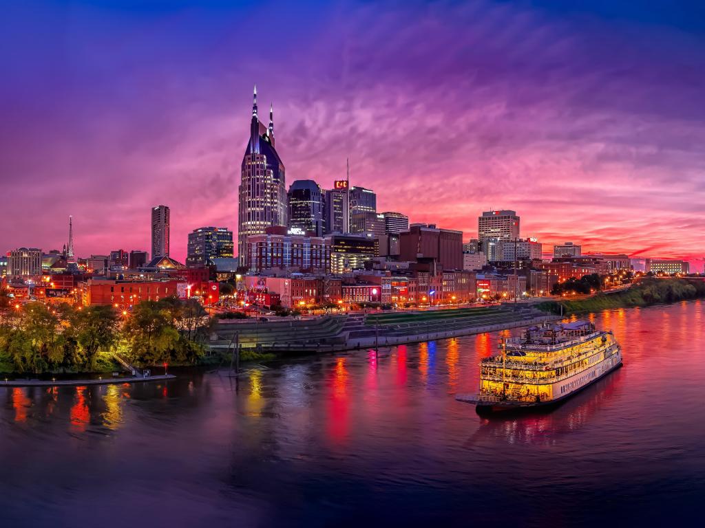 Illuminated river boat cruises past high rise buildings on the river bank, with deep pink and blue sunset light