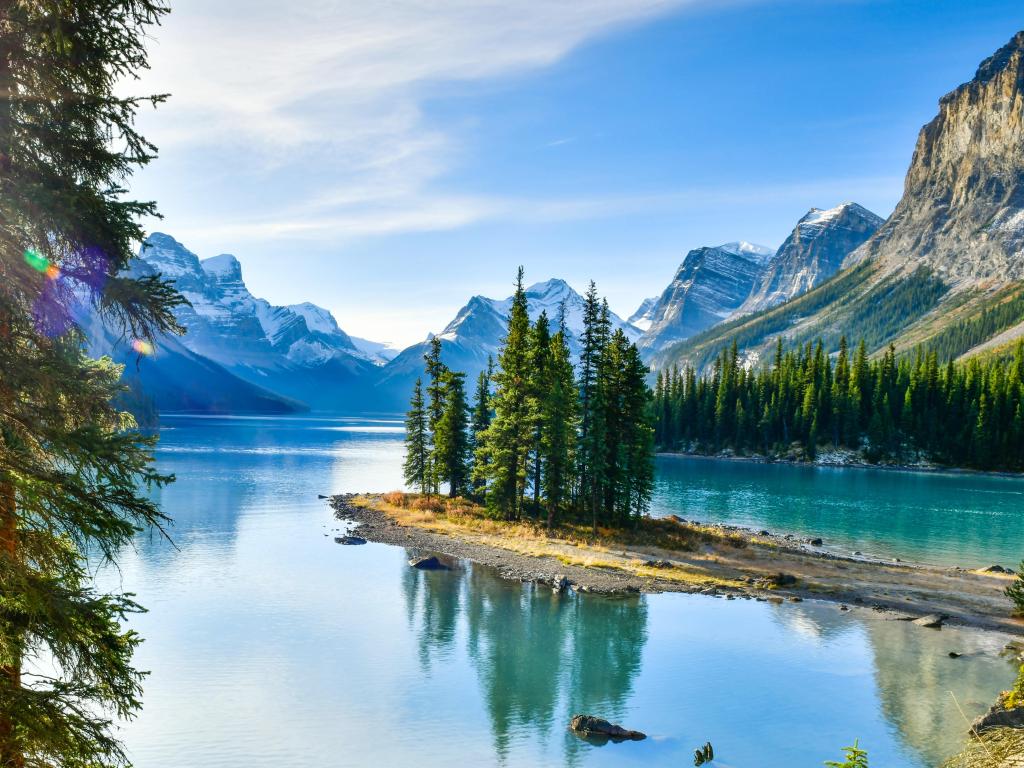 Panorama view of the sun beaming through the trees at Lake Maligne and mountain peaks in the background  