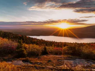 Gentle hill slopes covered in forest in the foreground, with sun setting over the water behind