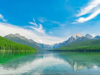 Glacier National Park, Montana, USA with a closeup of the Bowman Lake with trees and mountains in the distance against a blue sky.