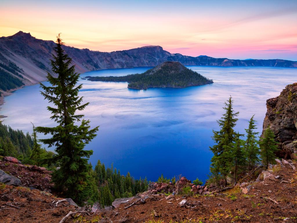 Crater Lake National Park, Oregon, USA taken at Wizard Island at sunset with trees in the foreground, a sunny lake and hills in the distance. 