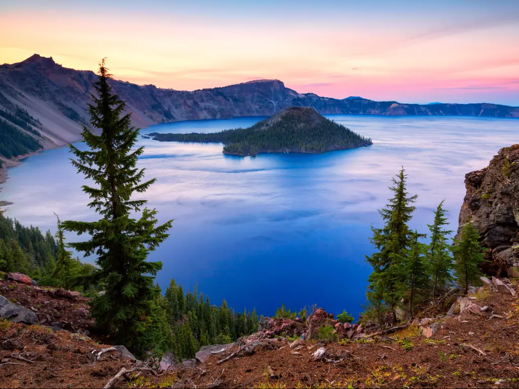 Crater Lake National Park, Oregon, USA taken at Wizard Island at sunset with trees in the foreground, a sunny lake and hills in the distance. 