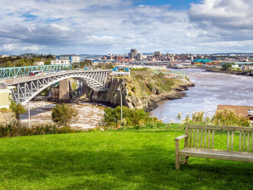 Saint John, New Brunswick, Canada from the Reversing Falls Park with an empty wooden bench in the foreground and a bridge leading towards the city in the distance. 