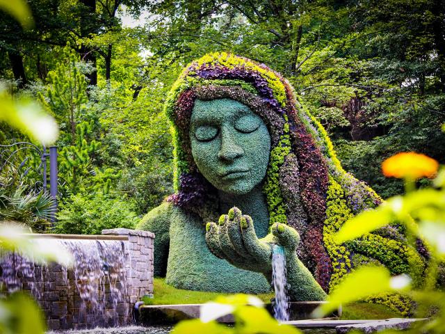 Earth goddess plant sculpture in the Atlanta Botanical Gardens. The bust is covered in grass while her hair is made up of flowers. Water is flowing from her palm. Her eyes are closed.