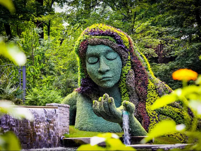 Earth goddess plant sculpture in the Atlanta Botanical Gardens. The bust is covered in grass while her hair is made up of flowers. Water is flowing from her palm. Her eyes are closed.