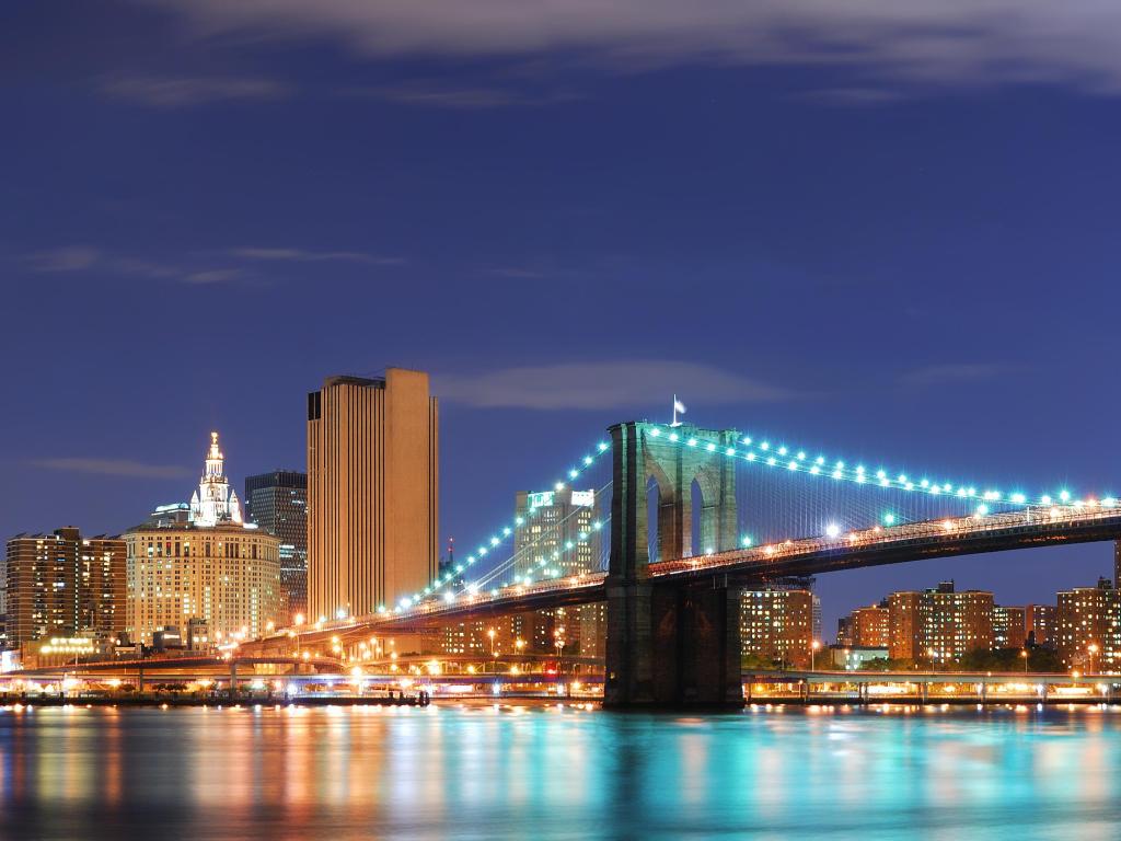 View of high rise buildings and illuminated Brooklyn Bridge at night, from across the river