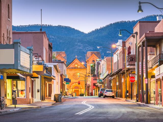 An image of Santa Fe, New Mexico during dusk with street lights on, buildings on both sides of the road, and the mountain in the background still visible.