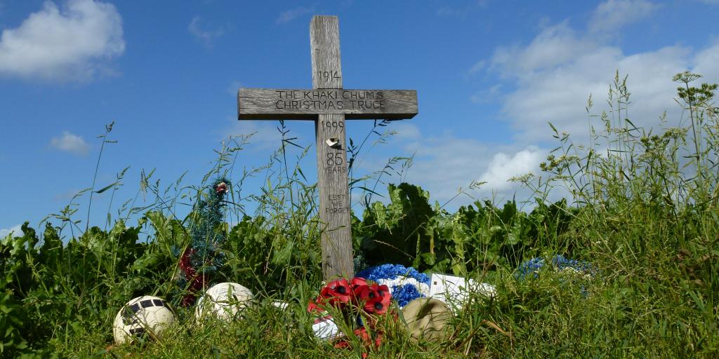 A cross in long grass surrounded by poppies and footballs at the Christmas Truce Memorial