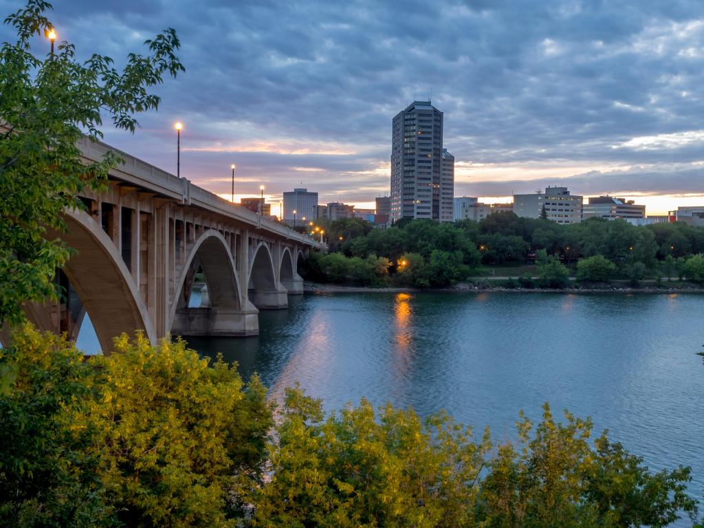 Saskatoon, Canada with the city skyline at night along the Saskatchewan River and a bridge in the foreground with trees surrounding it.