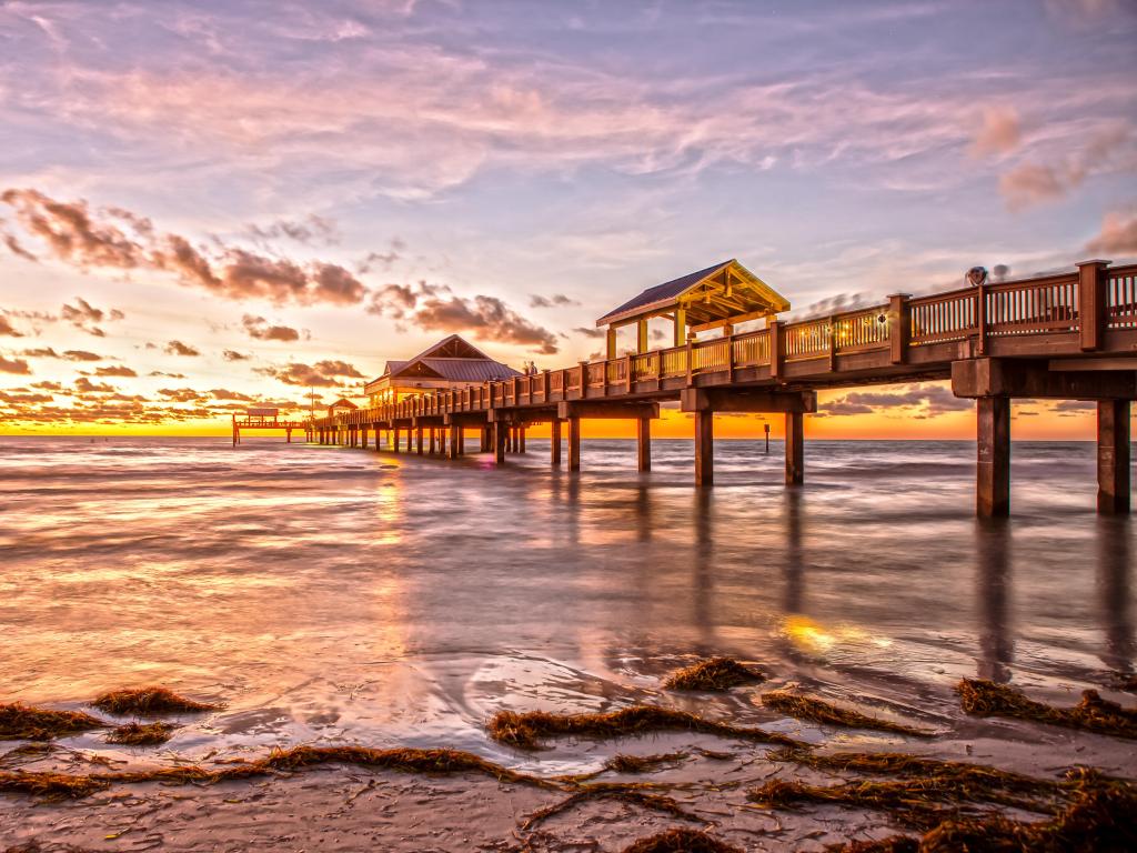 Long wooden pier stretches out into calm sea with pink, blue and gold sunset light reflected