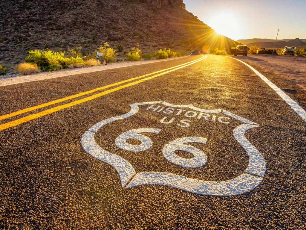 Route 66 road marking in white on black asphalt. A double yellow line runs through the centre off the road which disappears into the distance and the sunset.