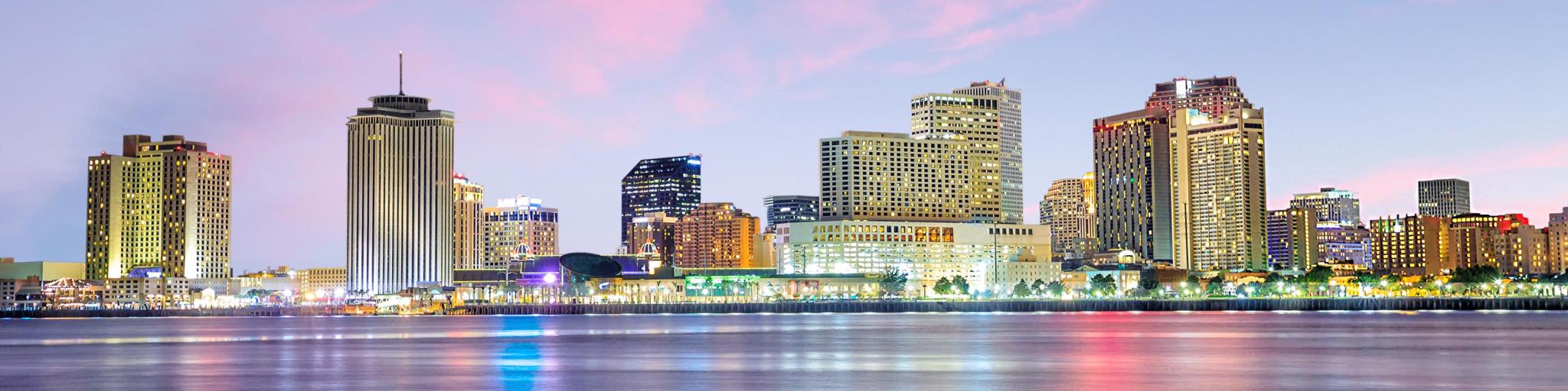 New Orleans, Louisiana with the downtown city in the background and the Mississippi River in the foreground at twilight under a pink sky.
