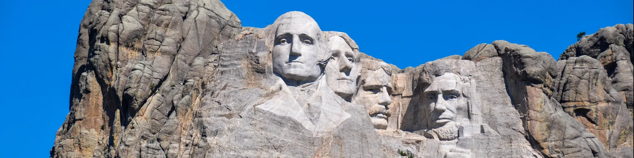 A scenic view of the 60-ft high granite sculpture of the four presidents of the United States in the Black Hills with a green forest at the foot of the mountain in a sunny morning in Mount Rushmore National Memorial Monument in South Dakota