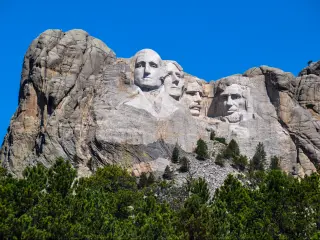 A scenic view of the 60-ft high granite sculpture of the four presidents of the United States in the Black Hills with a green forest at the foot of the mountain in a sunny morning in Mount Rushmore National Memorial Monument in South Dakota