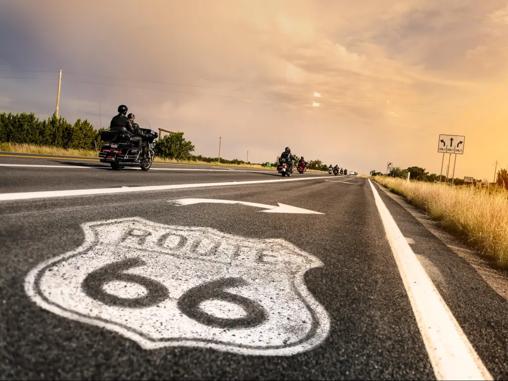 Route 66 roadsign painted onto the road with bikers passing, with bright sunlight illuminating the road and grass.