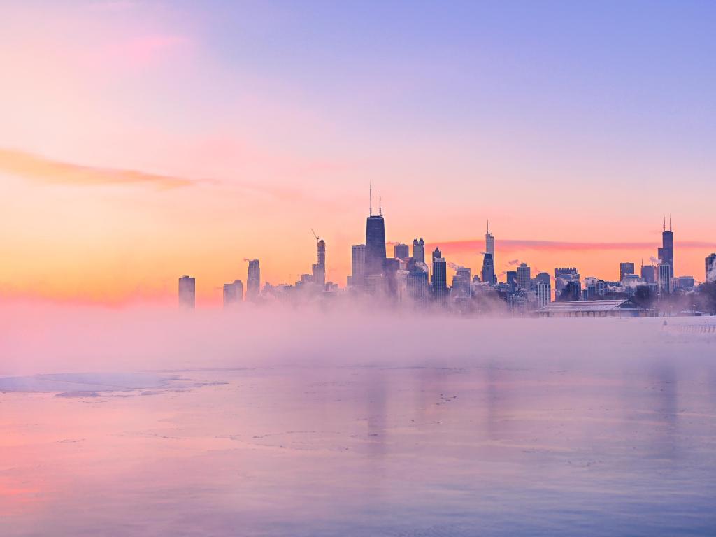 Pink sunrise light illuminates high rise buildings on the lake shore, with mist rising off the lake which is frozen over