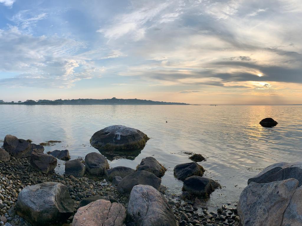 Views of Long Island Sound with pebbled shoreline and clear, calm waters in the forefront and cloudy sunset skies in the background