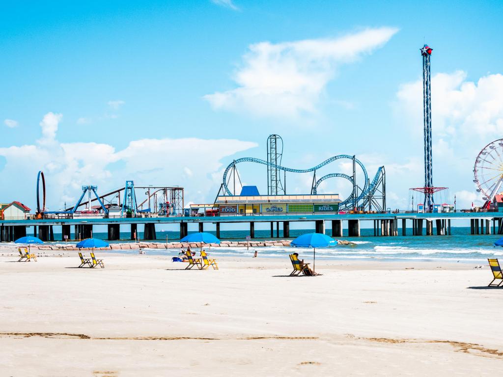 Galveston, Texas, USA with a view of the beach umbrellas and Pleasure Pier amusement park in the distance against a blue sky. 