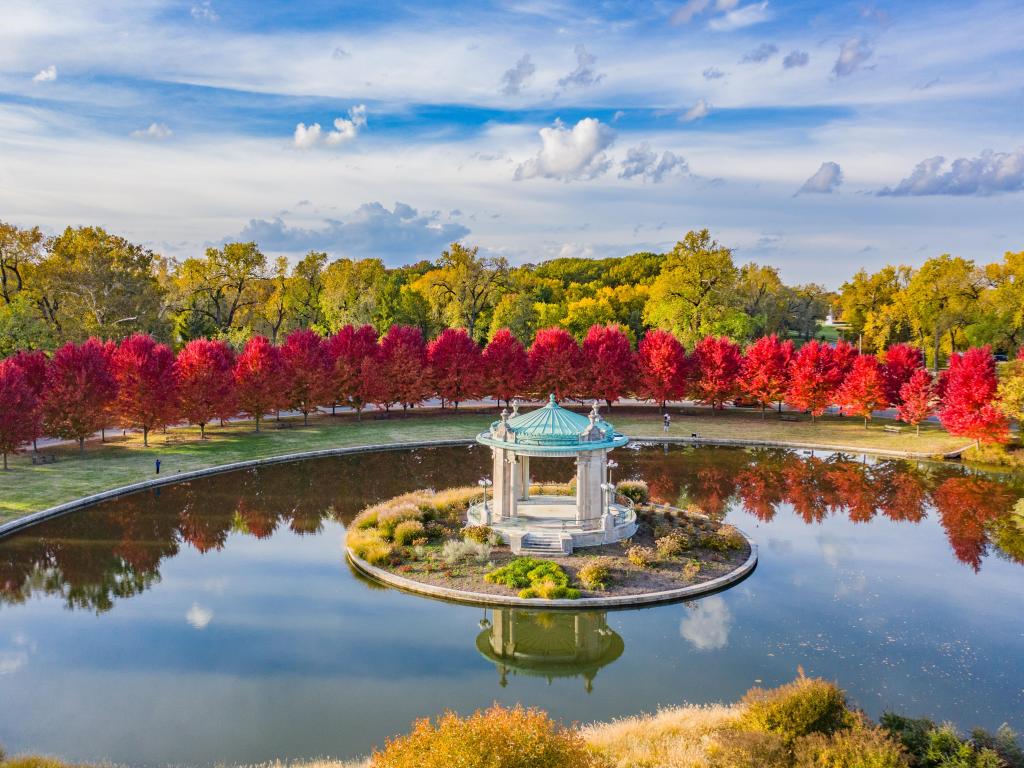 Forest Park bandstand in St. Louis. There is a row of autumnal trees in the background, surrounded by taller trees. The sky is cloudy with blue pathces.