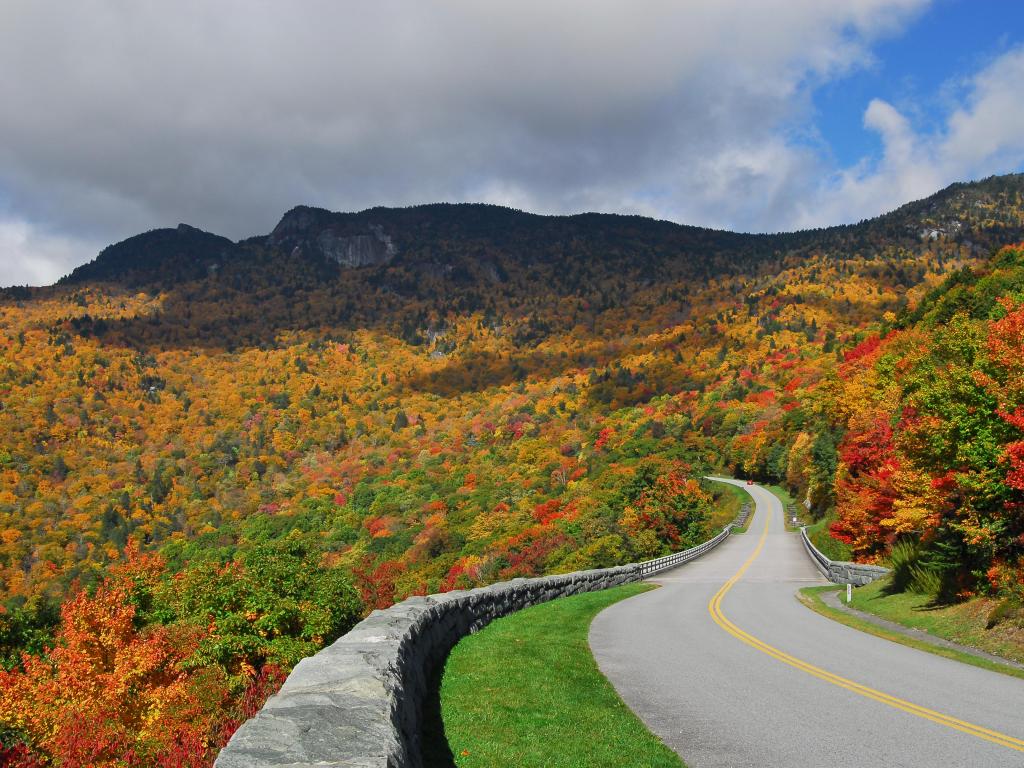 Blue Ridge Parkway, USA with a view of the Blue Ridge Mountains in the distance under a cloudy but blue sky, with the road leading towards trees at fall.