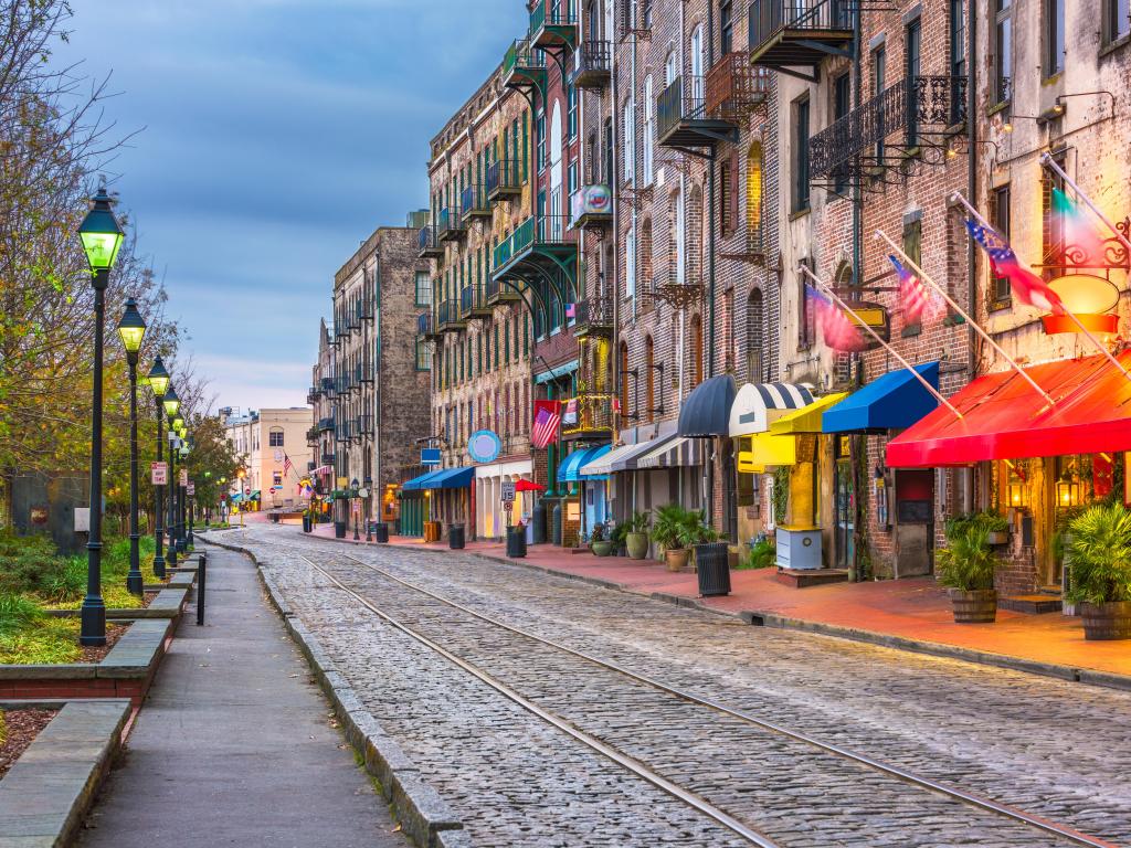 Savannah, Georgia, USA with a row of bars and restaurants on River Street at twilight with street lights and trees on the other side.