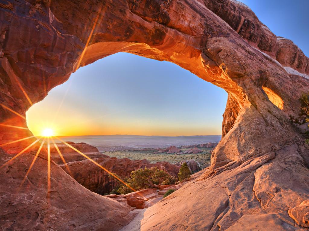 View through natural red rock arch at sunrise, across wide landscape with rocks and trees