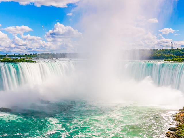 A panoramic view of the horseshoe-shaped falls with turquoise water the stunning Niagara Falls framed with green grasses and trees at summer in a blue sky with cotton-like clouds.