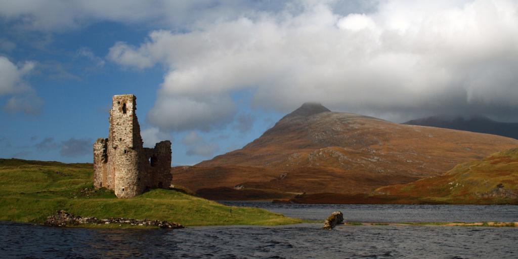 The ruins of Ardvreck Castle in Scotland with Loch Assynt surrounding it and a fiery mountain in the background