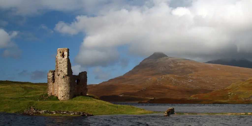 The ruins of Ardvreck Castle in Scotland with Loch Assynt surrounding it and a fiery mountain in the background