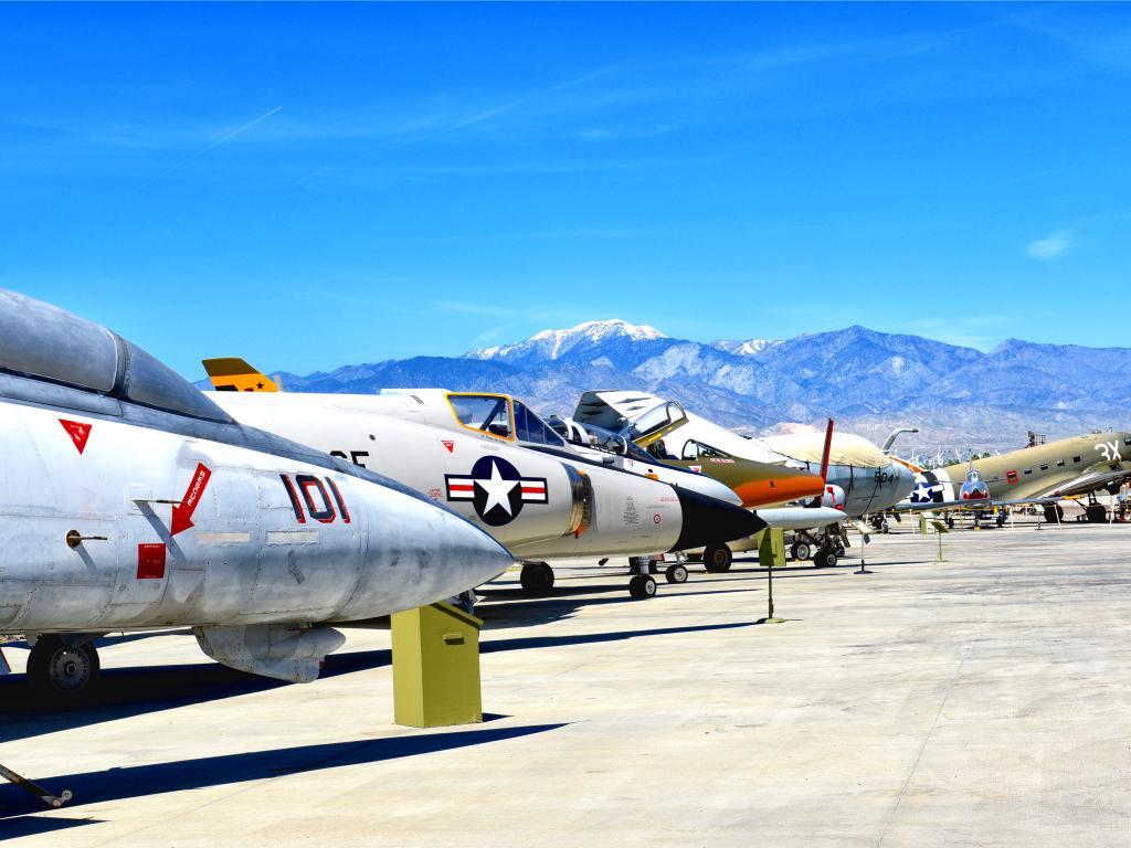 A line of vintage flyable WWII aircraft displays outside on a sunny day with a view of the mountains from afar at Palm Springs Air Museum.